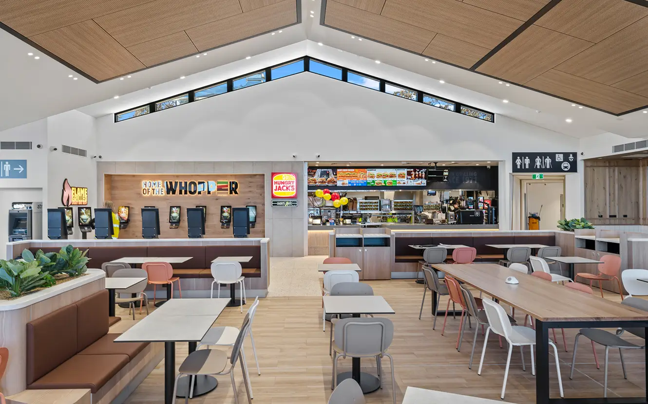 Modern fast food restaurant interior with wooden floors and ceilings. Tables and chairs are neatly arranged. A counter in the back displays menus and colorful signage. Self-service kiosks are on the left. Balloons add a festive touch.