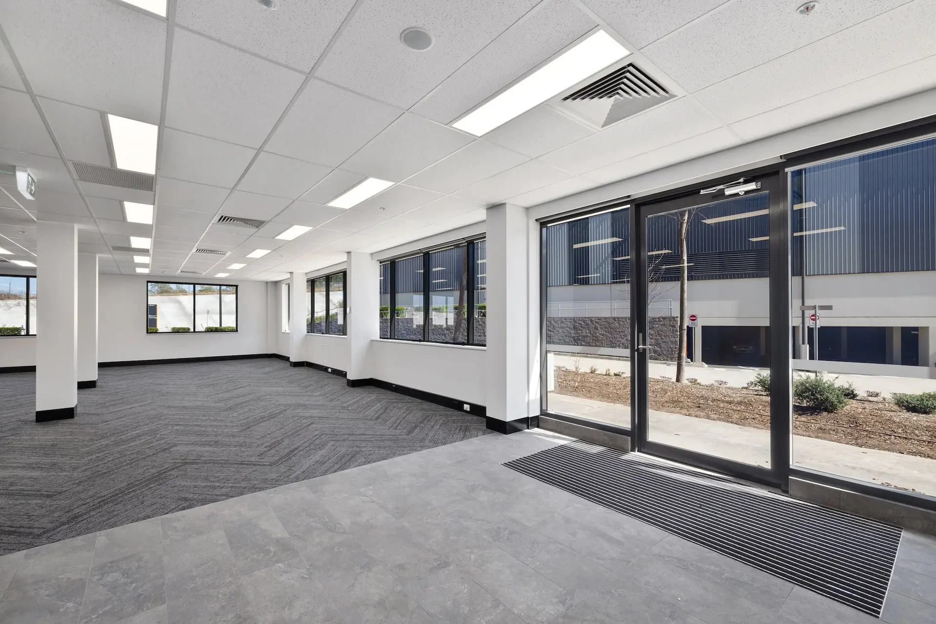 Spacious, modern office interior with large windows, grey carpet, and tile flooring. Multiple ceiling lights and visible exit signs enhance the bright, open atmosphere. Large glass doors lead outside to a building and landscaped area.