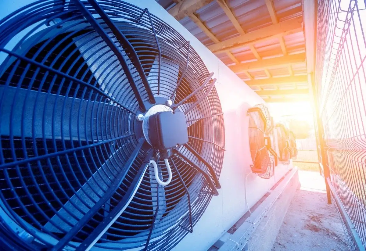 Industrial cooling fans are mounted in a row outside a building, partially shaded by a corrugated roof. The sun is setting on the right, casting a warm glow over the scene. A metal fence is visible on the right side.