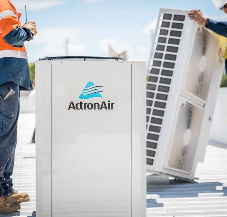 Two workers are installing an ActronAir HVAC unit on a rooftop. One holds a tool, while the other stands near the unit. The sky is clear and blue in the background.