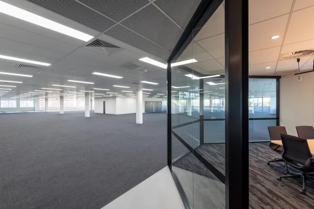 A spacious, empty office with gray carpet and fluorescent lighting. Large windows provide natural light. A glass-walled meeting room with chairs is visible to the right. The area is open, with a modern and minimalist design.