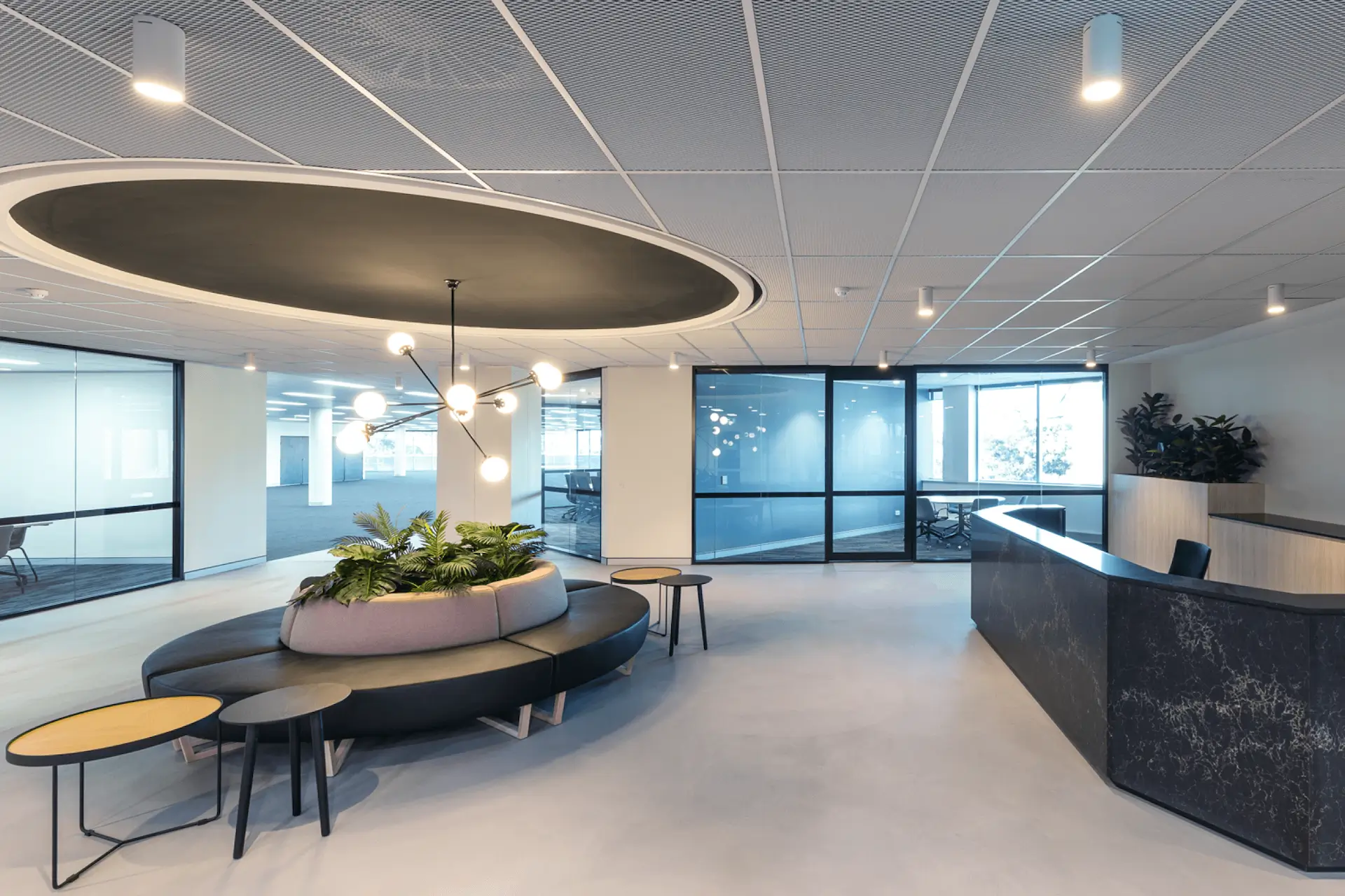 Modern office lobby with a round sofa and plant centerpiece beneath a circular ceiling feature. Surrounded by small tables, the area faces a sleek reception desk. Glass walls reveal a meeting room, and overhead lights provide illumination.