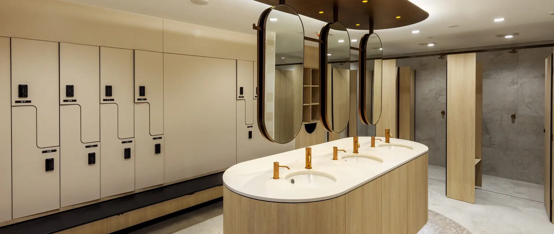 Modern locker room with minimalist design, featuring beige lockers, a curved bench, and an oval sink countertop with gold faucets. The room is well-lit with large, oval mirrors overhead and cubicles in the background.