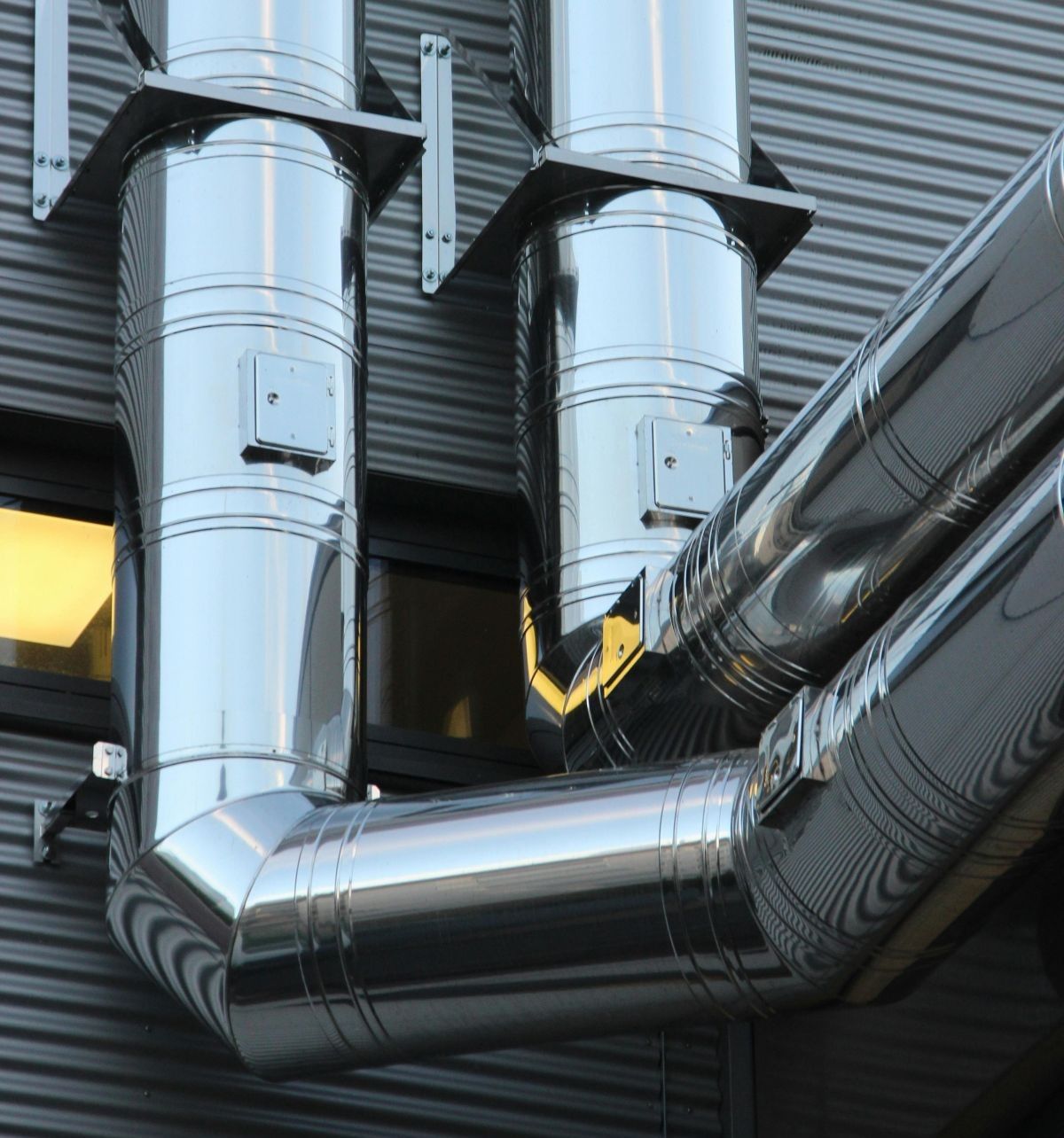 Close-up of shiny metal ventilation ducts mounted on a building with corrugated metal siding. Two rectangular windows with yellow light are visible behind the ducts.